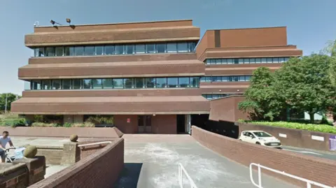 A multi-storey red-brick building with large glass windows and an entrance to a car park at the front of the building.