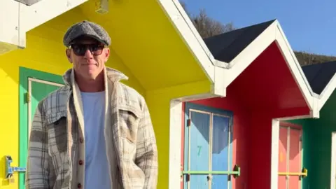 Luke Evans/Instagram Evans strands in front of colourful beach huts 