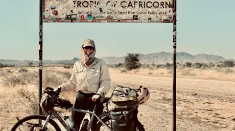 Ellie Mitchell-Heggs Mitchell-Heggs standing next to her bike with a Tropic of Capricorn sign behind her. She is wearing a sandy coloured hat with side panels and sunglasses and smiling at the camera. She has her bike next to her and is holding the handlebar and saddle. Behind her is a lot of sand and sandy foliage.