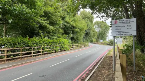 A view of the road up to the Warburton toll bridge. On the right hand side of the image there is a sign that says "bridge charge beyond this point" and it says that is free for motorcycles and £1 for other vehicles. It also says "pay online by midnight tomorrow night".