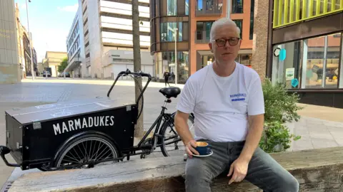 A man with grey hair and glasses is wearing a white t-shirt and grey jeans. He is sitting on a wooden bench with a coffee cup balance on his knee. In the background is an old-fashioned grocer's bike with the word Marmadukes and some city centre shops