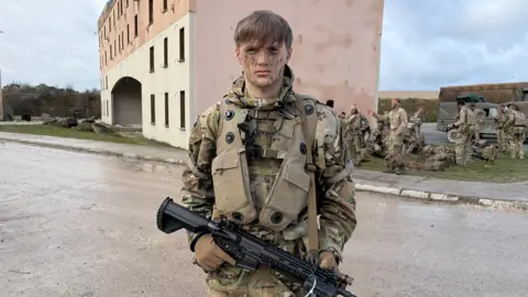 A man in army combat uniform holds a black gun over his body. He has camoflage paint on his face and army kit on his back. Behind him are more soldiers on the right of a pink and cream building. 