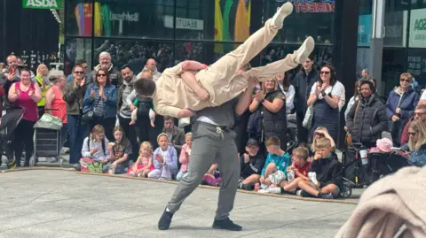 Male dancer in grey outfit lifts another man in a cream outfit in a performance in the town square. Children and adults are lined behind watching and applauding.