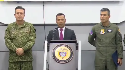Colombian Defence Minister Pedro Sánchez speaks at a news conference. He is flanked by two high-ranking members of Colombia's military. The minister is standing at a podium bearing the Ministry of Defence's official seal. 