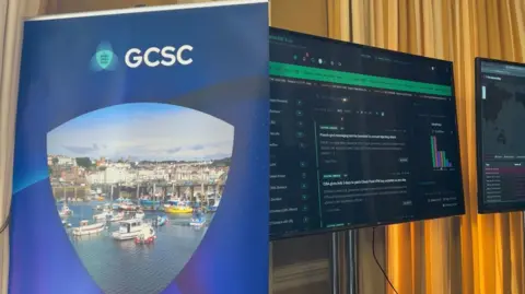 Two large black monitors on a yellow curtained wall show live tracking of cyber threats, including a map of the world. In front a blue banner which reads 'GCSC', with a photo St Peter Port Harbour 