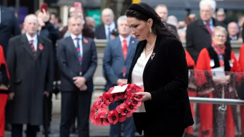 Pacemaker Emma Little Pengelly is holding a poppy wreath. Her head is bent. Crowds of people are watching.