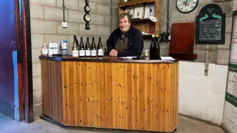 George Bowden, a 78-year-old man with dark hair wearing a navy fleece and brown jumper, sits behind a pine-clad bar in his winery in Leeds. There are five bottles of wine next to him and a barometer and a chalk board on the wall behind him.