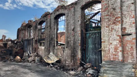 The derelict Salthouse Mills site in Barrow. A red brick wall with black scorch marks is surrounded by debris from a fire. The wall has arches where iron bars would have once been. There is no roof on the building.