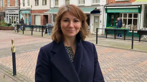 A woman standing on a pedestrianised street with shops in the background. She has brown hair and is wearing a dark coat. 