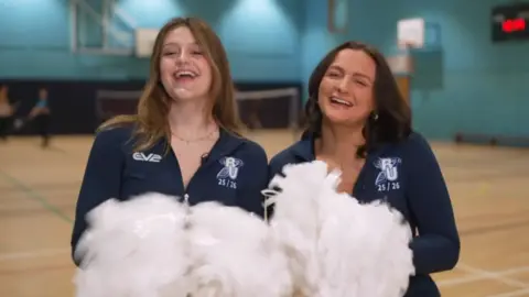 Eve Kimberley (left) and Liv Teague are wearing dark blue cheerleaders outfits and waving pompoms in a gym.