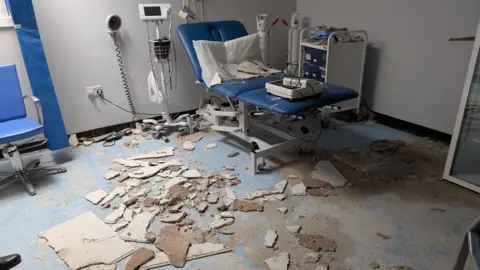 A view of the damaged room in the surgery. The roof has collapsed and parts of it is on the floor. There is a blue bed on the right as well as other equipment. 