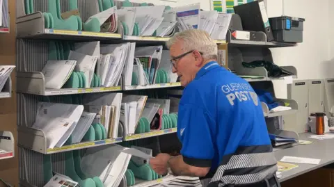 The image shows a the back of a man with white hair and spectacles wearing a blue polo shirt, sorting letters on a shelf.