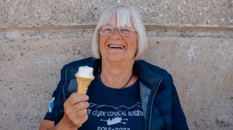 Kirstin McEwan A woman with grey hair and glasses in a blue T-Shirt and top smiles while eating an ice cream cone