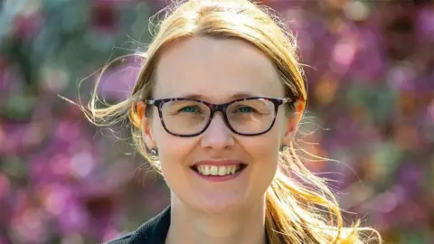 A headshot of Cat Smith MP, a young woman with brown hear, smiling for a camera wearing brown tortoise shell glasses. 