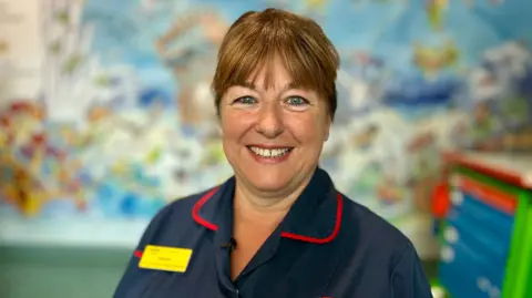 Sarah is wearing her dark blue nurse's uniform, trimmed with red piping around the collar. Her chestnut brown hair is tied back with a fringe just covering her eyebrows. She is smiling at the camera. In the background is a brightly coloured cartoon mural covering the wall of one of the wards in Picu.
