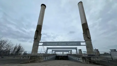 The entrance to the National Glass Centre. The concrete path leads down a ramp. Meanwhile, two concrete pillars border either side. A grey sign reads: "National Glass Centre".
