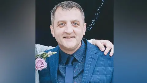 Head and shoulders shot of David Dosa. He is wearing a blue checked jacket, blue shirt and tie. He has an arm around his shoulders and the owner of the arm has a pink flower on his grey jacket, although we cannot see the rest of him.