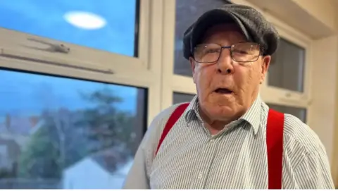 A man wearing a white striped shirt with red braces, glasses and a flat cap stands in front of a large window inside a building. 