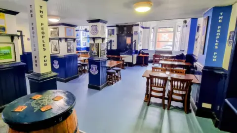 The inside of The Winslow Hotel pub. The floor is light blue and there are wooden tables and chairs. There is a lot of Everton FC memorabilia and slogans on the walls.