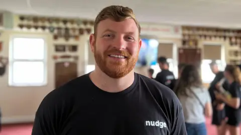 Tim is wearing a black T-shirt with the word “nudge” stands in the foreground of a clubhouse. Several people are gathered in the background, which is softly blurred. The room has red carpet, large windows, display plaques on the walls, and a television screen in the distance.