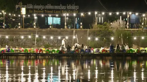 A string of lights illuminates the river. In the bank, people are sitting on brightly colored chairs. 