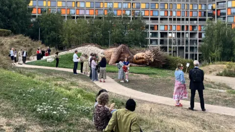 BBC/Kat Cowen A large wooden sculpture lies on the grass in-front of a huge colourful block of flats. A path winds through a grassy area in front of the sculpture and people sit around the park. 
