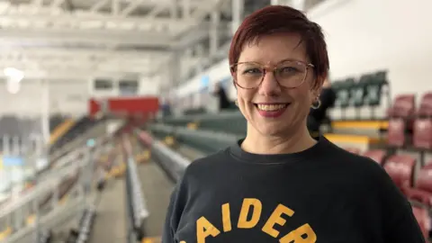 Ice hockey fan Emily Garside standing in the stand of an ice hockey arena. She has short, swept over hair that is reddish brown and is wearing circular tortoise shell framed glasses. Emily is smiling and wearing a black jumper that says 'Raiders' in bright yellow above a shield logo with the letter R in the style of a sports team