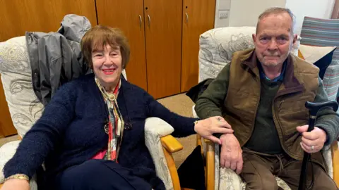 Woman with short brown hair, navy cardigan and patterend scarf sat next to a man, holding his hand. The elderly man is holding a walking stick and wearing a green sleeveless jacket with a dark green jumper underneath