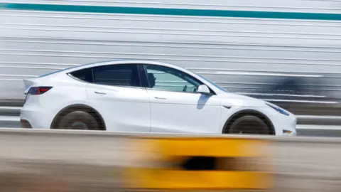 A white car drives on a road, blurred in the background. A man is visible behind the wheel.