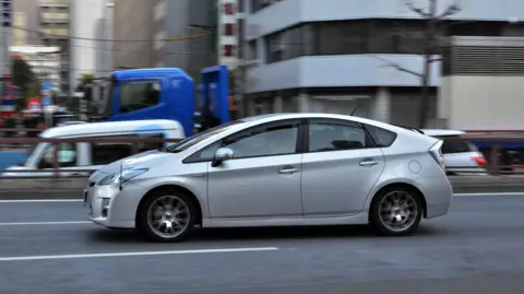 Getty Images A silver Toyota Prius is pictured driving down an urban road from the side. 