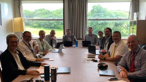 A group of people all sat around a long rectangular table, with notebooks or laptops in front of them. There are two large windows at the back of the room with greenery visible through them.