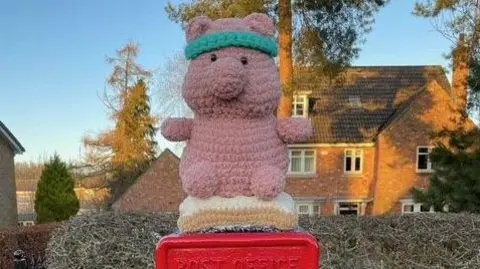 The crocheted pig sits on a blanket attached to a post box in Brough.  A pretty cottage is in the background.