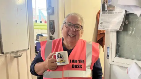 Karen Davies, a woman with pulled back grey hair, black rimmed glasses is smiling and holding a mug with a picture of Wonder Woman. Karen is wearing a peach flourescent vest with the words 'I am the boss' on her chest
