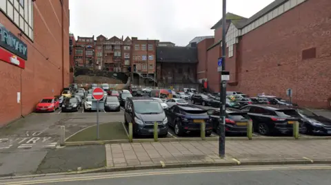 The Gap car park, Shrewsbury, with cars parked in the spaces, No Entry signs in the foreground, near a pavement and a road with double yellow lines. There is a lamp post with signs attached to it on the pavement and there are town buildings in the background.