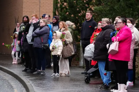 PA Media Several adults and children, some holding flowers, stand on a pavement watching a funeral procession. Many are wearing boots and heavy winter jackets.