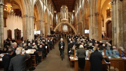 Athena Rows of mourners seated inside Llandaff Cathedral. They are facing towards the alter, with their backs towards the camera