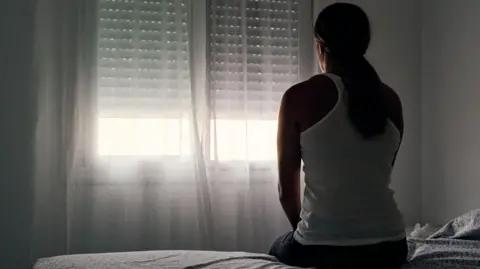 A woman has her back to the camera and is sitting on the edge of a bed in a dark room, facing the window. She has her hair tied back into a ponytail and is wearing jeans and a white tank top.