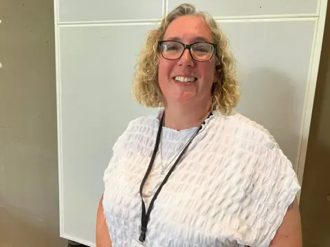 BBC/Amy Holmes Hayley Whitaker, who has curly blonde hair, glasses and is wearing a white top, smiles at the camera. She is wearing a lanyard.