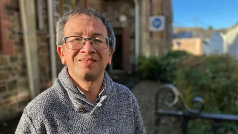 A man with short grey hair wearing rectangular glasses wearing a grey jumper standing outside a brown building.