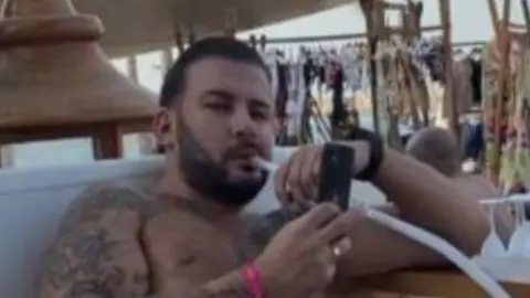 James Harding smoking a cigaratte and looking at his phone with his top off