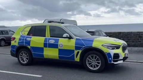 BBC A picture of a Guernsey police car. It is taken from the side of the vehicle and the logo is visible. There are two cars parked facing a brick wall. The sea is beind the wall. The sky is grey and filled with clouds. 