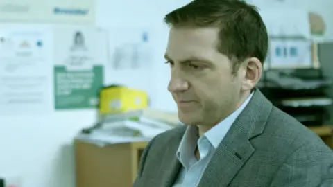 Dr Faisal Maassarani, wearing a pale blue shirt and grey plaid suit jacket, is sitting at a computer in his surgery in an image from a 2018 BBC health documentary. 