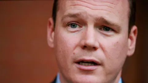 A man speaking to the camera. The image is a close up in high focus. The man has blue eyes and short dark hair.