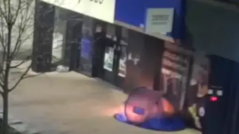 A blue tent is seen shining as it has been set on fire outside a Tesco Express on a high street. The CCTV image is blurry.