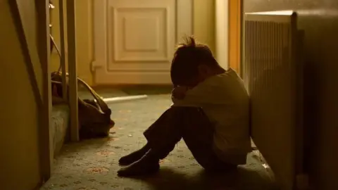 A young child sits alone on a patterned carpet in a dimly lit hallway, leaning against a radiator. Their posture is withdrawn—head bowed and resting on their knees, arms wrapped around their legs. A closed door is in the background, and a small bag with scattered belongings lies nearby.