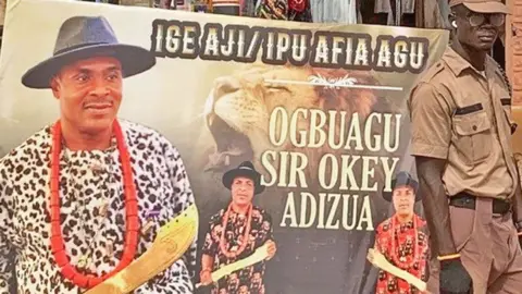 A uniformed security officer stands to the right of a poster advertising a ceremony for a man called Sir Okey Adizua to become a 'Leopard Slayer'. There are three images of a man holding a gold sword, wearing a large red bead necklace and a wide-brimmed black hat