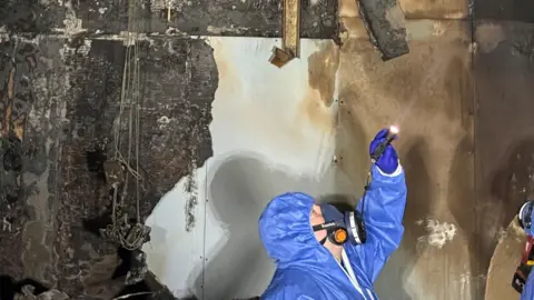 University Hospital Southampton A man in blue overalls and a protective mask shines a torch up towards the ceiling as he inspects fire damage at the hospital. 