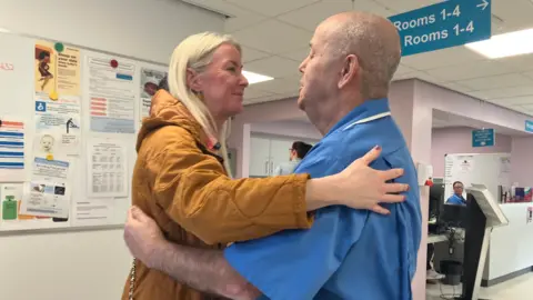 Laura Rooney, a woman with long, straight blonde hair, leans in to hug Fraser Morton, a man with shaved grey hair, in a hospital corridor. She is wearing a tan-coloured quilted overcoat and carrying a handbag with a gold and black chain. He is wearing a blue midwife's tunic. They are both smiling.