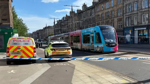 A city street scene cordoned off by emergency services on a sunny day. Blue-and-white police tape stretches across the foreground, blocking the road. Behind the tape, a marked police van with high-visibility red and yellow chevrons and the word “POLICE” is parked on the left, next to an ambulance response car with yellow and green markings. A modern tram in bright blue and pink livery is stopped on tracks running through the middle of the street.
The street is lined with multi-storey stone buildings featuring rows of windows, chimneys, and ground-floor shops, including signs such as “The Leith Walk Gallery” and “Greet the Ch…” (partially obscured). Overhead tram wires and poles run along the street. A few pedestrians are visible on the pavement behind the cordon, and some people can be seen further up the road near the emergency vehicles.
The sky is clear blue with scattered clouds, and strong sunlight casts long shadows across the pavement and road surface. Yellow road markings and tactile paving are visible in the foreground near the blocked crossing. The scene suggests an active incident with the area restricted for safety.