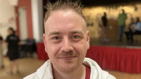 Richard Prior smiling in front of a stage. He has short spikey blonde hair.